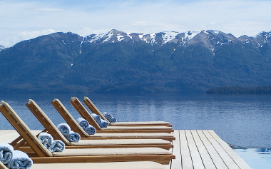 A row of wooden lounge chairs with rolled-up towels sit on a deck overlooking a calm lake. Snow-capped mountains rise in the background under a partly cloudy sky, creating a serene and picturesque setting.