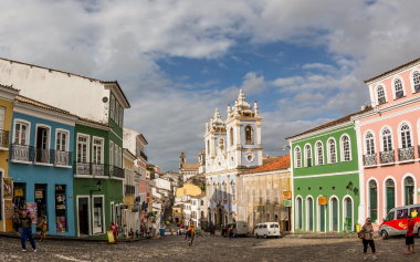 A vibrant street scene in a historic town with colorful buildings lining a cobblestone street. A church with two towers stands prominently in the background under a partly cloudy sky. People are walking and a few cars are parked along the street.