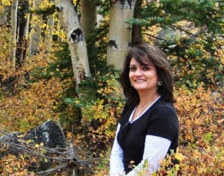 A woman with long, dark hair is smiling at the camera while standing in a lush forest with green and yellow foliage. She is wearing a black top over a white long-sleeved shirt, and behind her are trees and bushes with autumn leaves.