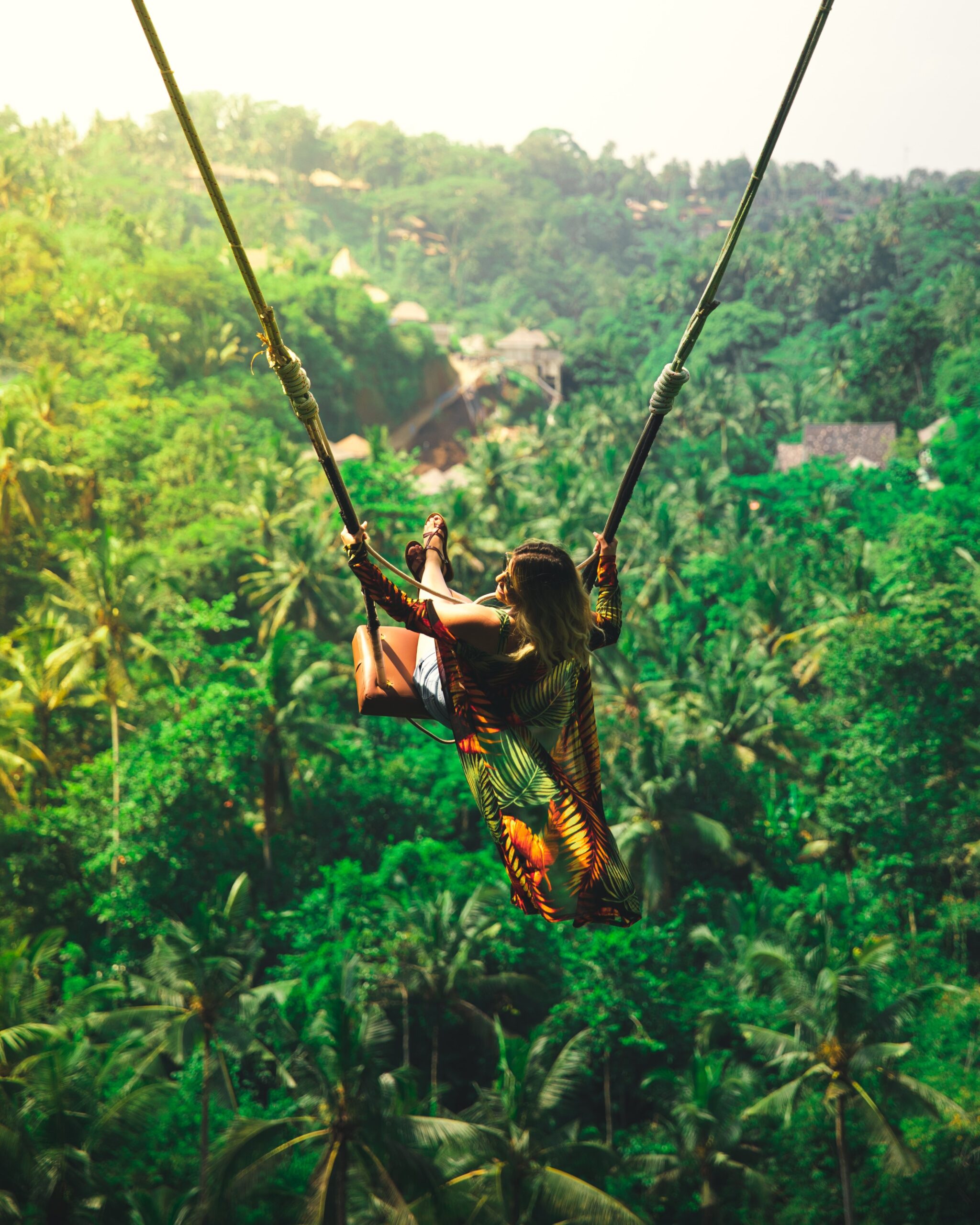 A person in a colorful outfit swings high above a lush, green tropical forest, with sunlight filtering through the trees and a few buildings partially visible in the background.