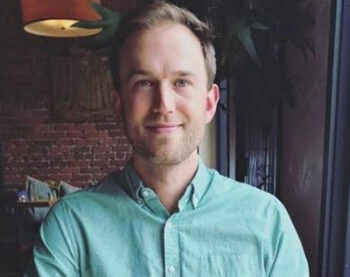 A man with short hair and a beard, wearing a light green button-up shirt, sits in a dimly lit room with an exposed brick wall in the background. He is smiling slightly and looking at the camera.