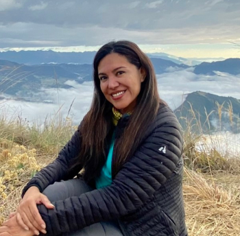 A woman with long dark hair is sitting on a grassy hillside overlooking a scenic vista of mountains and clouds. She is wearing a black jacket and smiling at the camera, enjoying the natural landscape in the background.