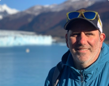 A man wearing a blue jacket, yellow sunglasses, and a baseball cap smiles at the camera. Behind him is a scenic view of a glacier with mountains and a body of water in the background, bathed in sunlight.