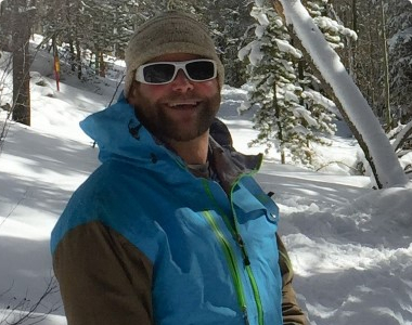 A person stands outdoors in a snowy forest, wearing a blue jacket, gray knitted hat, and white sunglasses. The individual is smiling, and the surroundings are filled with snow-covered trees.