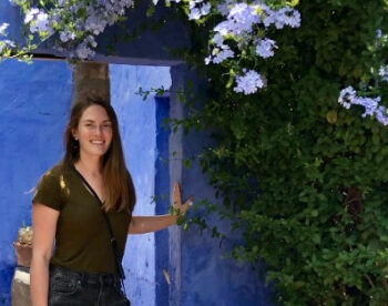 A woman with long brown hair wearing a green shirt and black shorts stands smiling in front of a vibrant blue wall with lush green foliage and blooming purple flowers overhead. She rests her hand on the doorway, with a crossbody bag slung over her shoulder.