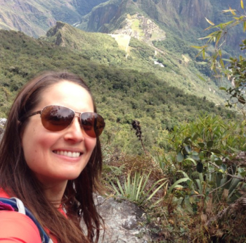 A woman with long brown hair, wearing aviator sunglasses and a red shirt, smiles for a selfie with a lush, green mountainous landscape in the background. The scene includes dense vegetation, steep slopes, and distant ruins.