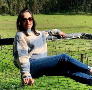 A person wearing sunglasses, a striped sweater, and jeans is lounging on a net-like structure in a green field with trees and grazing horses in the background. The person appears relaxed and is leaning back, enjoying the outdoor setting.