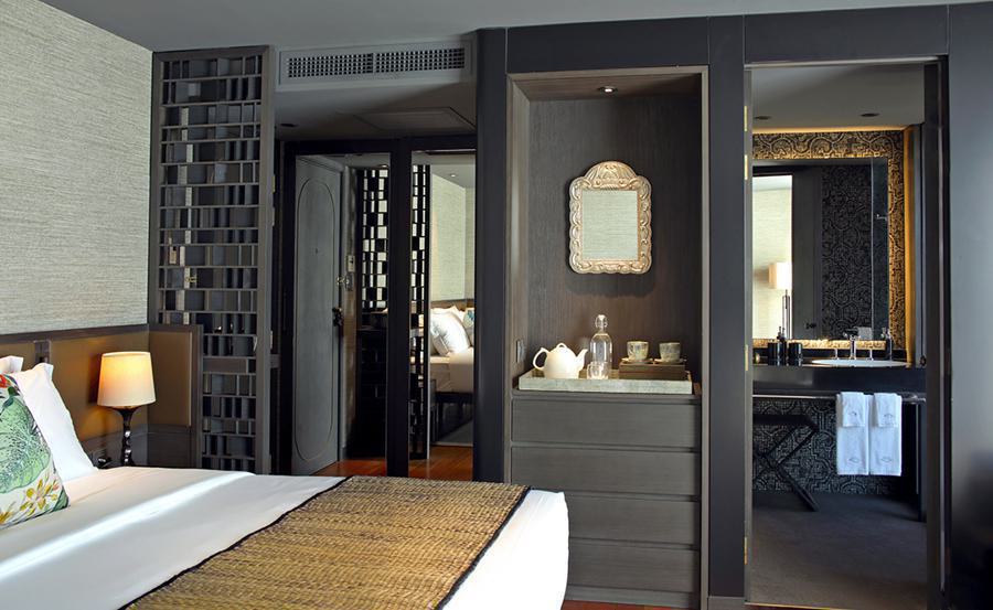 Modern hotel room featuring a bed with white linens and a floral pillow. A nightstand with a lamp is positioned next to the bed. There is a wooden partition, a small shelf with a tea set, and an open doorway leading to a sleek bathroom with towels and a mirror, reminiscent of Aqua Nera Amazon River cruise accommodations.