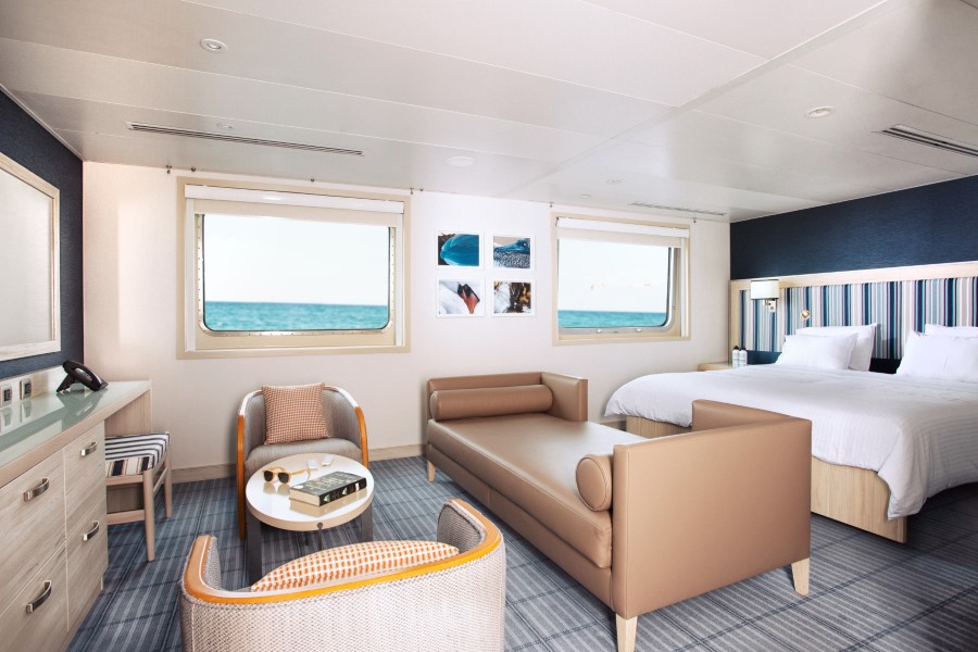 A spacious cruise ship cabin on the Santa Cruz II Galapagos cruise features large windows showing an ocean view. The room includes a double bed, a beige sofa, a small table with books and glasses, two chairs, nightstands, a dresser with a phone, and wall art. The decor is modern and light-colored.