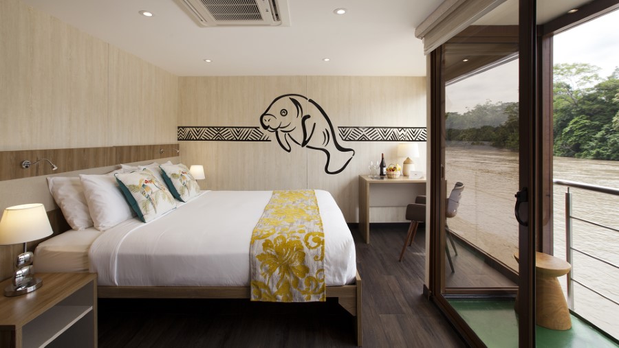 A modern, well-lit bedroom with a large bed adorned with white linens and yellow floral accents. A manatee mural decorates the wall above the bed, and the room boasts large windows with river views. There's also a small desk and a balcony with green flooring, perfect for imagining an Amazon River cruise.