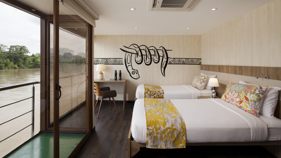 A well-lit room with two single beds adorned with floral-patterned bedding. A mural of a hand holding a snake decorates the wall behind the beds. There is a desk with a chair, and a sliding glass door opens to a balcony with a view, perfect for gazing after an Amazon River cruise.