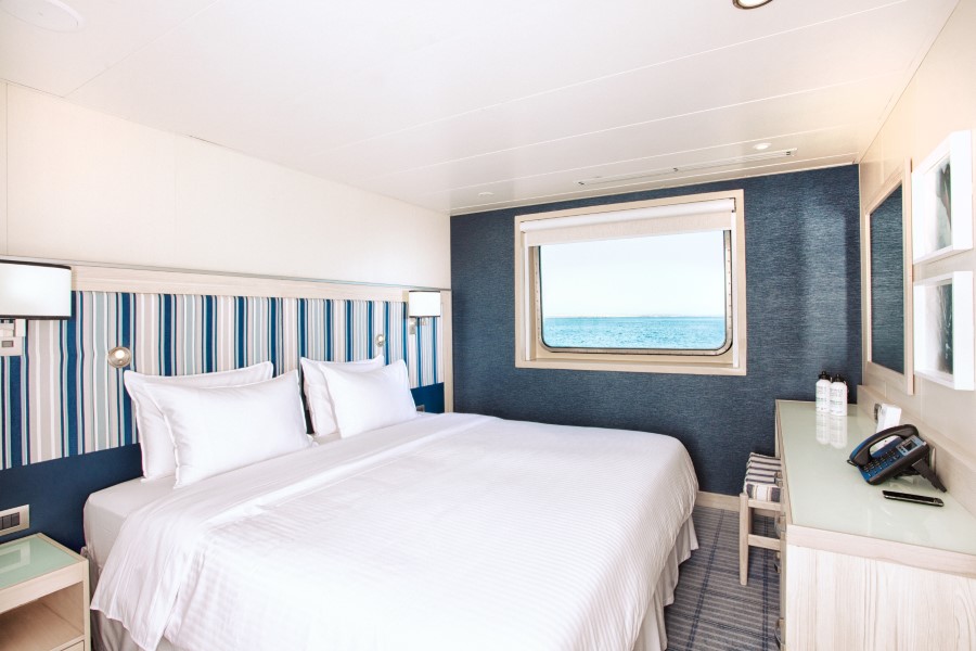 A bright, modern bedroom with a double bed dressed in white linens. The room features a navy and white striped accent wall and a large window offering an ocean view. A desk with water bottles, phone, and a stool is on the right. Inspired by the Santa Cruz II Galapagos cruise, the room is lit by natural light.