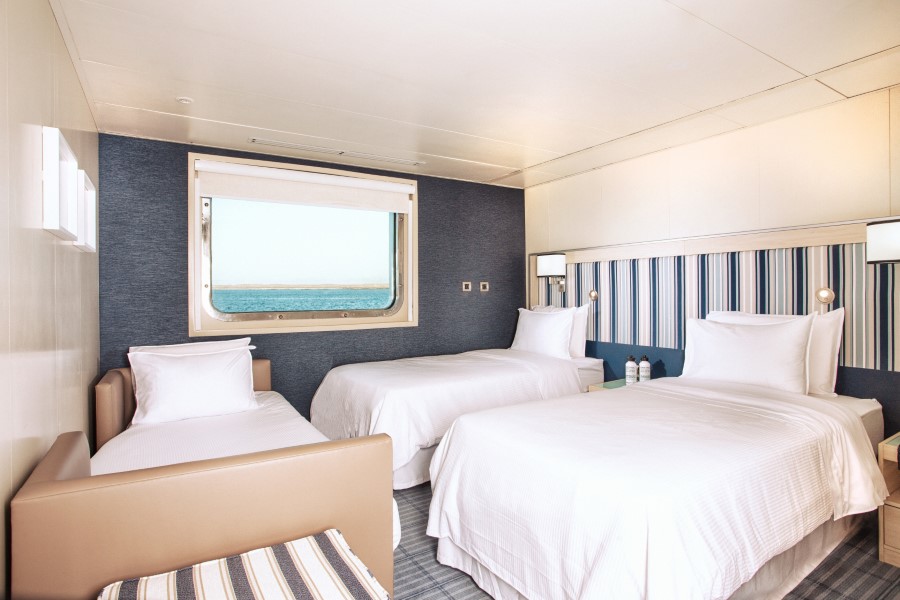 A bright, modern bedroom featuring two single beds with white linens, a striped headboard, a comfortable tan couch with a striped cushion, and a large window showing a view of the ocean. Perfect for relaxing after exploring on your Santa Cruz II Galapagos cruise, two water bottles are placed on a small nightstand between the beds.