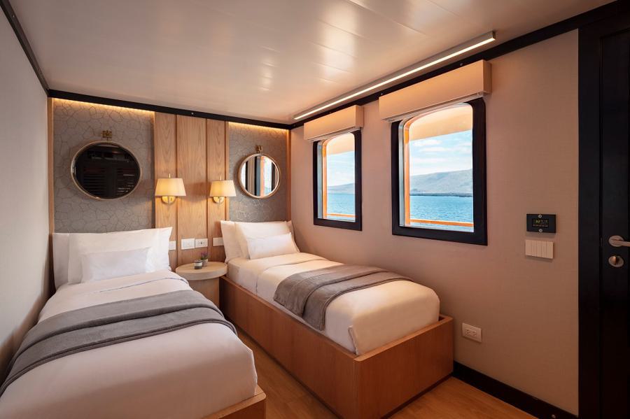 A cozy room with two single beds, each decorated with gray and white bedding. The room features two round mirrors, wall lamps, and two windows showing an ocean view. The walls are a mix of light-colored wood and neutral tones, and the wooden flooring adds to the charm. Perfect for a Grace Galapagos cruise experience.