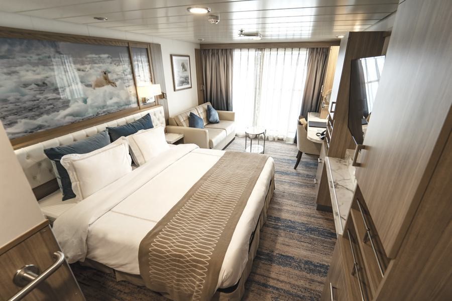 A well-appointed hotel room on the Greg Mortimer Antarctica expedition features a bed with white linens and decorative pillows, a seating area with a sofa, a desk and chair, a wall-mounted TV, and a large window with curtains offering an ocean view.