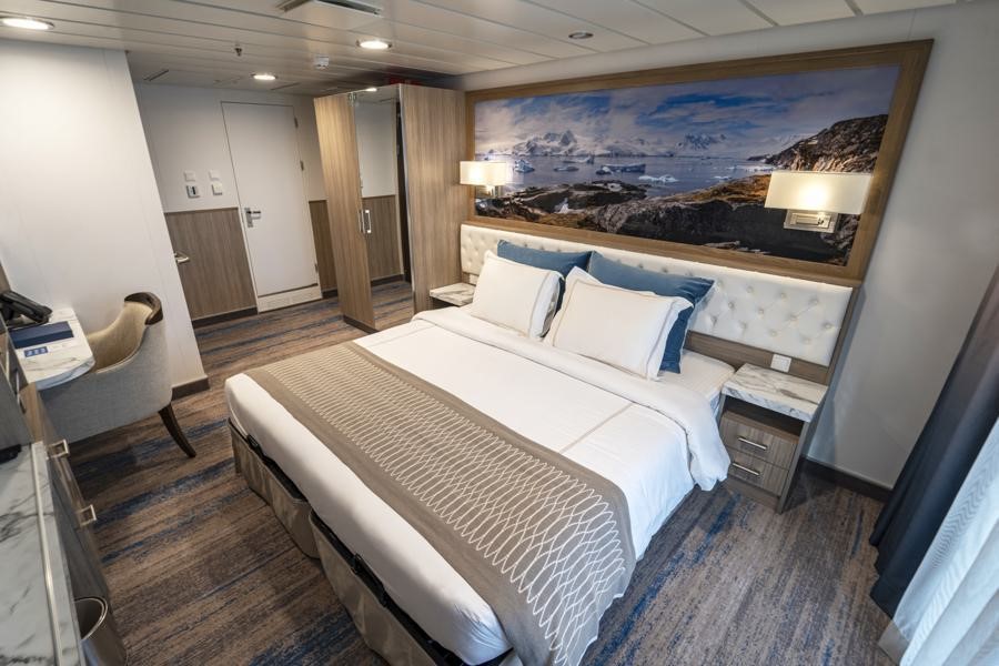 A modern, spacious hotel room with a large bed adorned with white and beige linens and blue accent pillows. The room features a wall-mounted landscape painting, a desk with a chair, bedside tables with lamps, and a window with curtains that fills the room with natural light. Perfect for guests embarking on the Greg Mortimer Antarctica expedition.