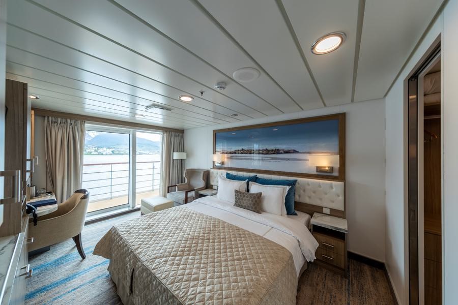 A well-lit, modern bedroom with a king-size bed adorned with white and beige linens. The room features a large window and a sliding glass door leading to a balcony with ocean views suitable for dreaming about a Greg Mortimer Antarctica expedition. There are two cushioned chairs, a wooden side table, and a built-in desk.