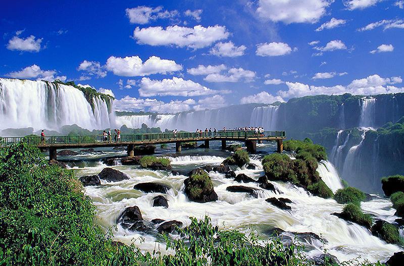 A group of people stands on a wooden platform overlooking a breathtaking view of Iguazu Falls surrounded by lush greenery. The sky is bright blue with scattered white clouds, enhancing the vibrant, natural beauty of the scene.