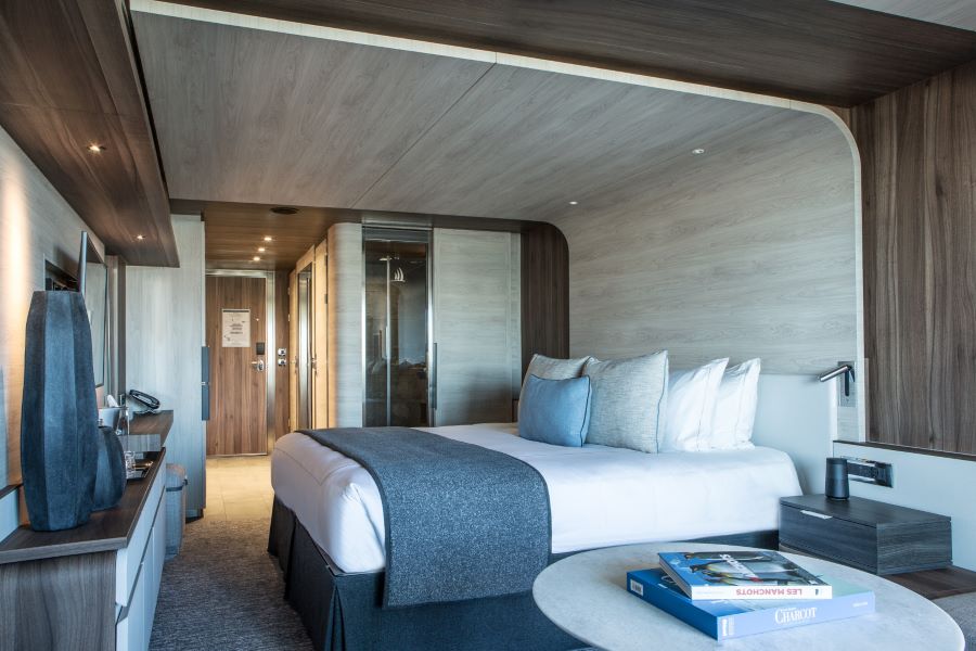 Modern hotel room with a neatly made bed featuring white linens and a gray throw blanket. The room has wooden accents, a flat-screen TV, a small round table with books, and a view of the hallway leading to a wooden door. A glass-walled shower is visible in the bathroom area.