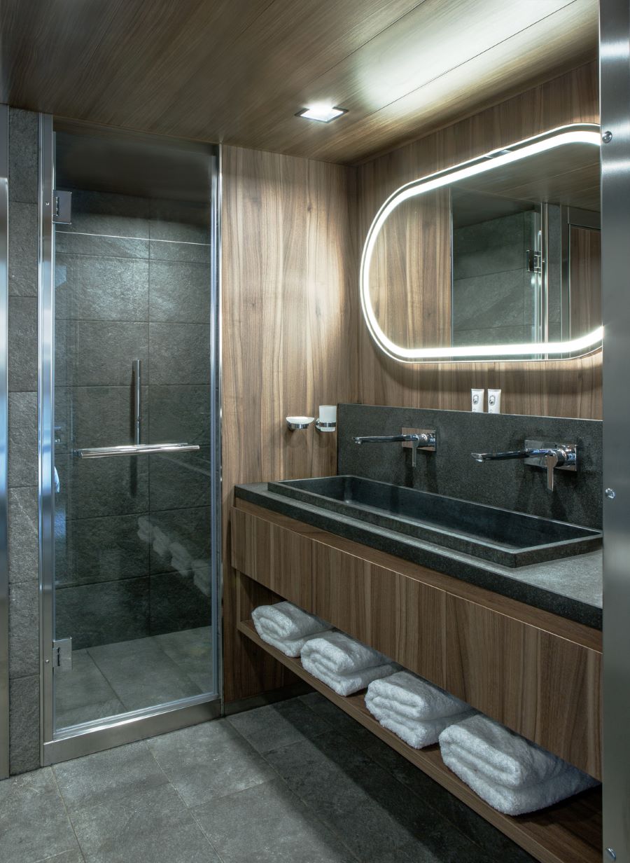 A modern bathroom features a glass-enclosed shower on the left with dark gray tiles. Next to it is a wooden vanity with a black countertop, two sinks, and wall-mounted faucets. An oval, backlit mirror hangs above, and neatly folded white towels are stored below.