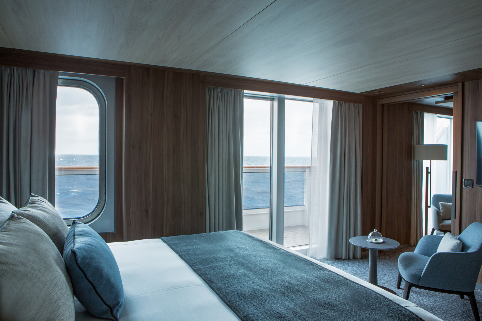 A cozy cruise ship cabin with a large bed, decorated in neutral tones and blue accents, featuring two large windows revealing an ocean view. The room has light wooden walls, floor-to-ceiling curtains, a small sitting area with two chairs, and a lamp.