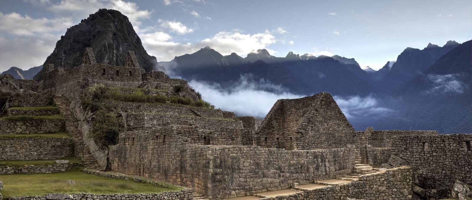 A scenic view of Machu Picchu, an ancient Incan city located in Peru. Stone ruins and terraced fields lay against a backdrop of misty mountains under a partly cloudy sky. The sunlight gently illuminates the stone structures, highlighting their intricate details.