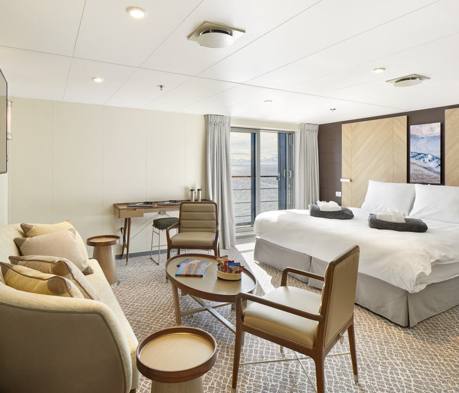 A luxurious and spacious hotel room features a large bed with white linens, a sitting area with a cream-colored sofa, two brown chairs, and a round coffee table. A desk and chair are positioned near the glass doors that open to a scenic ocean view, perfect for planning your Magellan Explorer Antarctica expedition.