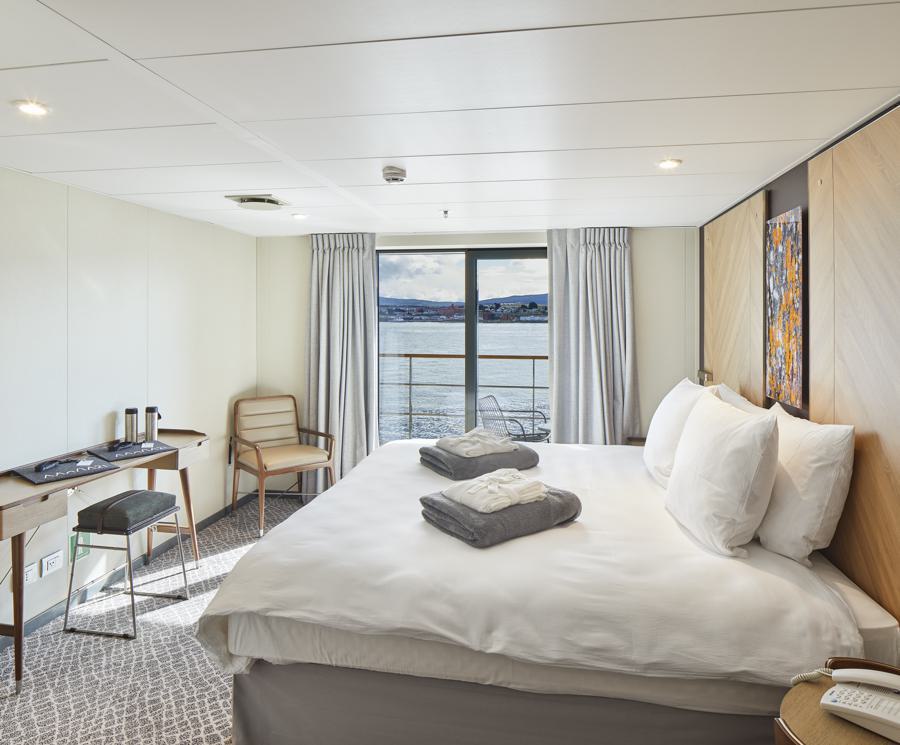 A modern, well-lit cruise ship cabin featuring a large bed with white linens and folded robes, a desk with a chair, a small table with another chair, and a window with a view of the sea and coastline. Light wood and neutral tones dominate the décor, perfect for guests on the Magellan Explorer Antarctica expedition.