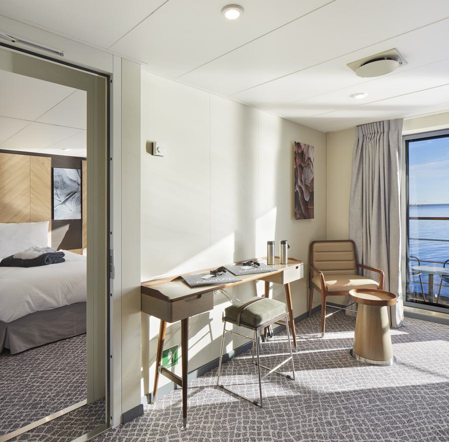 A bright, modern hotel room with a view of the sea. The room features a desk with a lamp, chair, and assorted items, a small seating area with two chairs and a round table, and an adjoining bedroom with a neatly made bed. Natural light fills the space through a large window, perfect for planning your next Magellan Explorer Antarctica expedition.