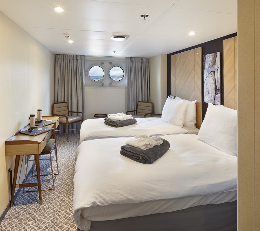 A cozy cruise ship cabin on the Magellan Explorer Antarctica expedition features two neatly made twin beds with folded towels and bathrobes on top. The room includes a desk, chairs, and two circular porthole windows letting in natural light. The decor boasts wooden accents and wall art.