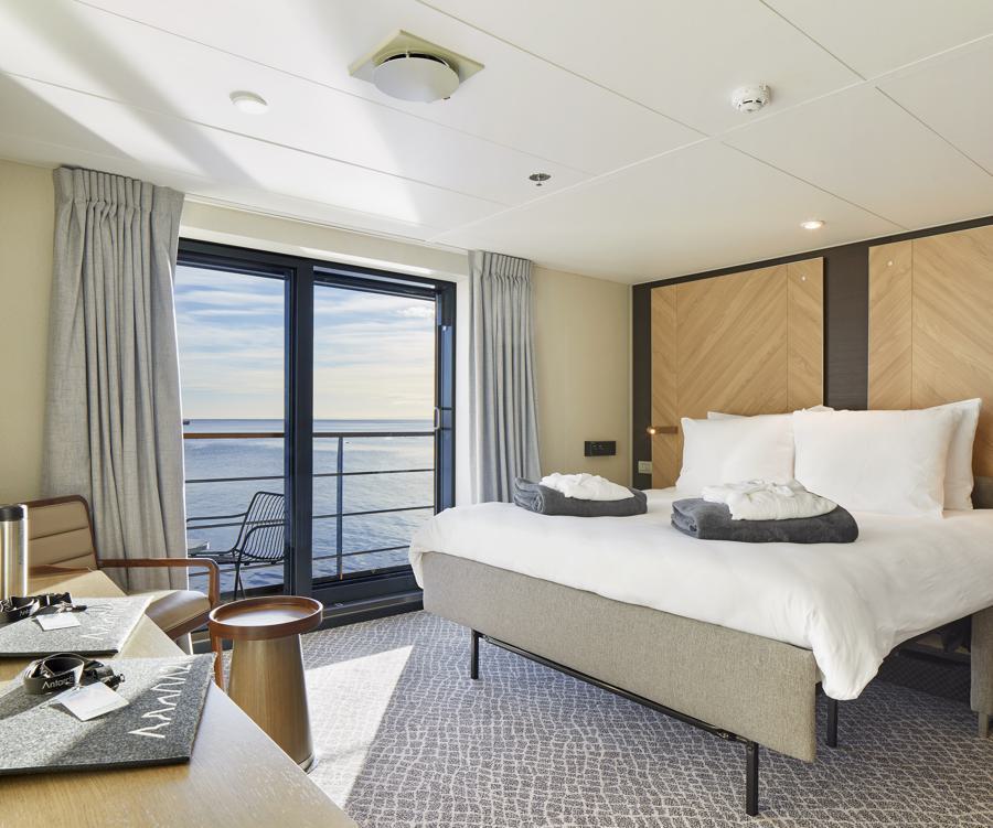 A modern hotel room with a large bed neatly made with white linens and robes folded at the foot. The room features a large window with a view of the ocean, a small table with two chairs, and light-colored decor with wooden accents, perfect for those inspired by the Magellan Explorer Antarctica expedition. Natural light fills the space.