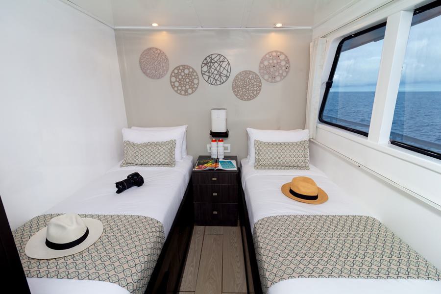 A cozy cabin on a Monserrat Galapagos cruise features two single beds adorned with patterned blankets and pillows. Between them is a nightstand with books, a lamp, and water bottles. Hats and a camera rest on the beds. A large window shows a view of the ocean. Textured wall art decorates the space.