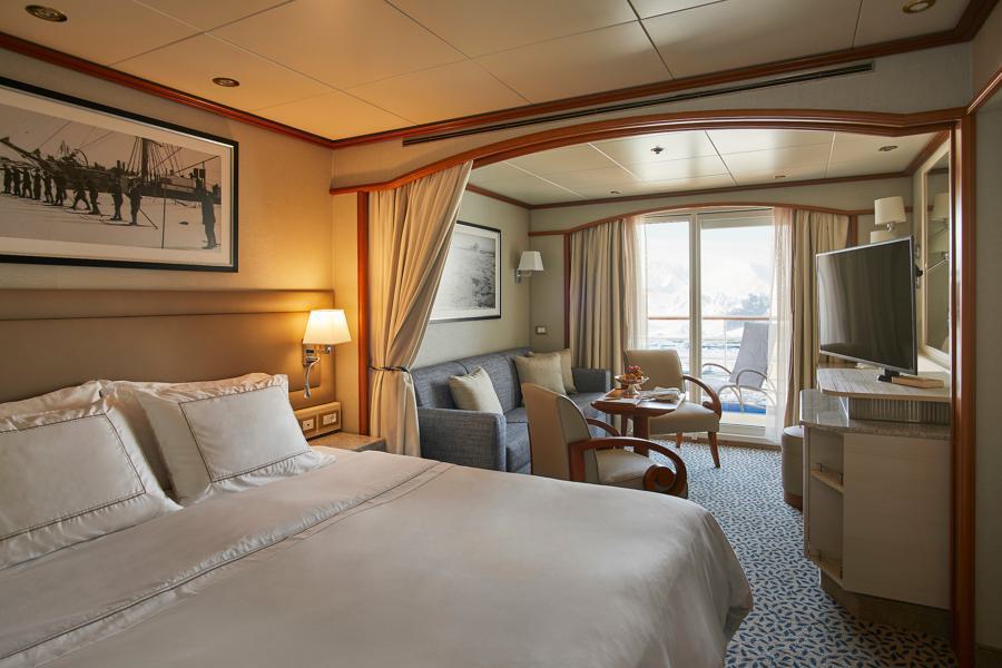 A well-lit cruise ship cabin with a double bed, a sofa and table, a flat-screen TV, and a private balcony with sliding glass doors. The room has elegant decor with framed artwork, light fixtures, and comfortable seating, offering a view of the ocean on your Silver Cloud Antarctica expedition.