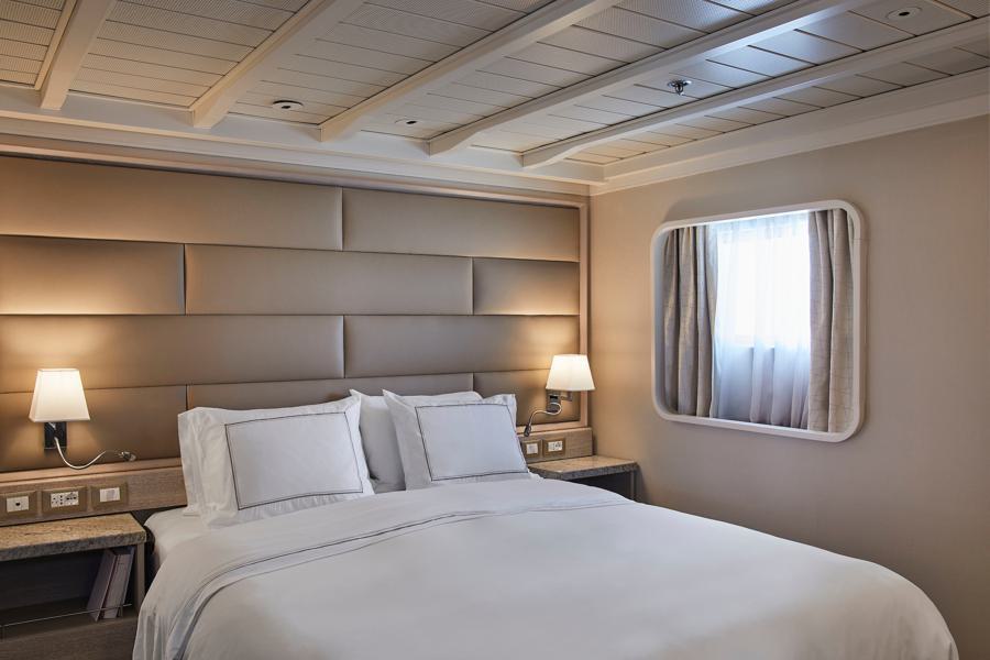 A cozy bedroom with a neatly made king-sized bed adorned with white linens and pillows, a modern taupe padded headboard, two bedside tables each with a lamp, and a window with a sheer curtain framed by a rectangular mirror. The ceiling features a wooden beam design that evokes the tranquility of a Silver Cloud Antarctica expedition.