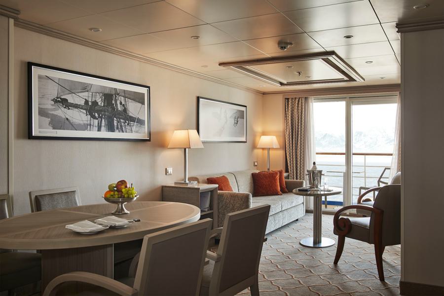 A spacious and elegant living area features a gray sofa, two armchairs, a round wooden dining table with four chairs, and two framed paintings on the wall. A bowl of fruit sits on the table, and floor-to-ceiling windows offer a view of snow-capped mountains, reminiscent of a Silver Cloud Antarctica expedition.