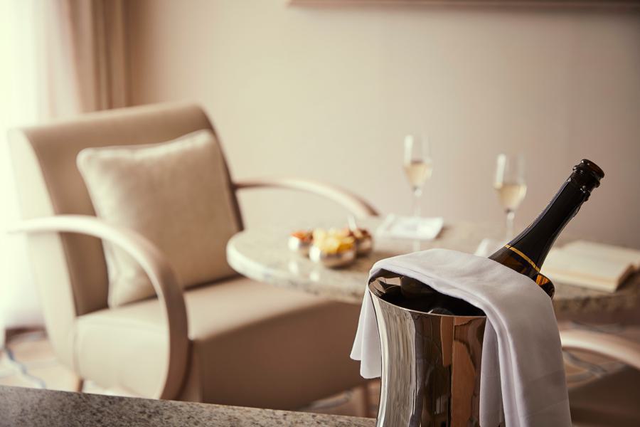 A chilled bottle of champagne in an ice bucket with a white cloth draped over it is positioned in the foreground. In the background, a round table with two champagne glasses, snacks, and an open book is set between two beige armchairs in a softly lit room, perfect for discussing your next Silver Cloud Antarctica expedition.