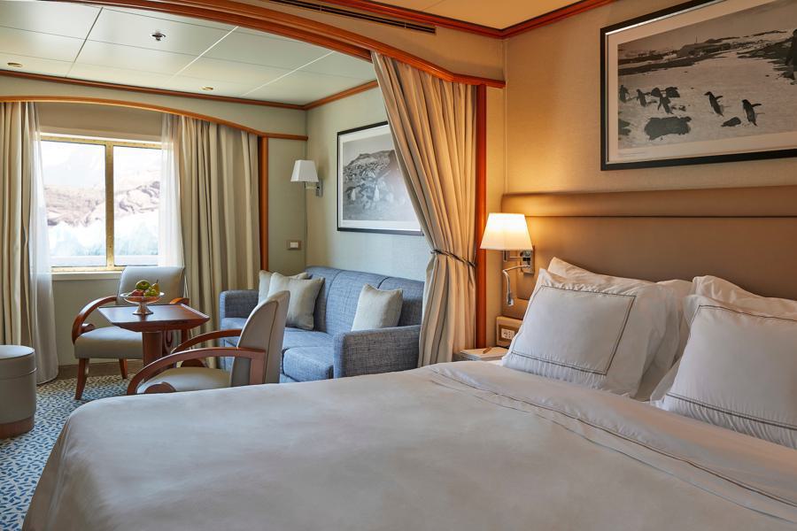 A cozy, well-lit hotel room features a neatly made bed with white linens, a sitting area with a blue sofa, a wooden table, and two chairs. A window with curtains allows natural light to stream in. Two framed black-and-white photographs decorate the walls, adding an air of Silver Cloud Antarctica expedition charm.