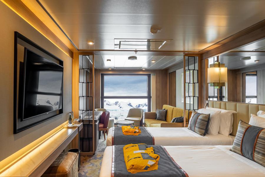 A well-appointed, modern cruise ship cabin with twin beds, each with a yellow life jacket on top. The room has a large TV, side table with water and glasses, couch, and large windows offering a scenic view of snowy mountains. Warm lighting enhances the cozy atmosphere onboard the World Explorer Antarctica expedition.