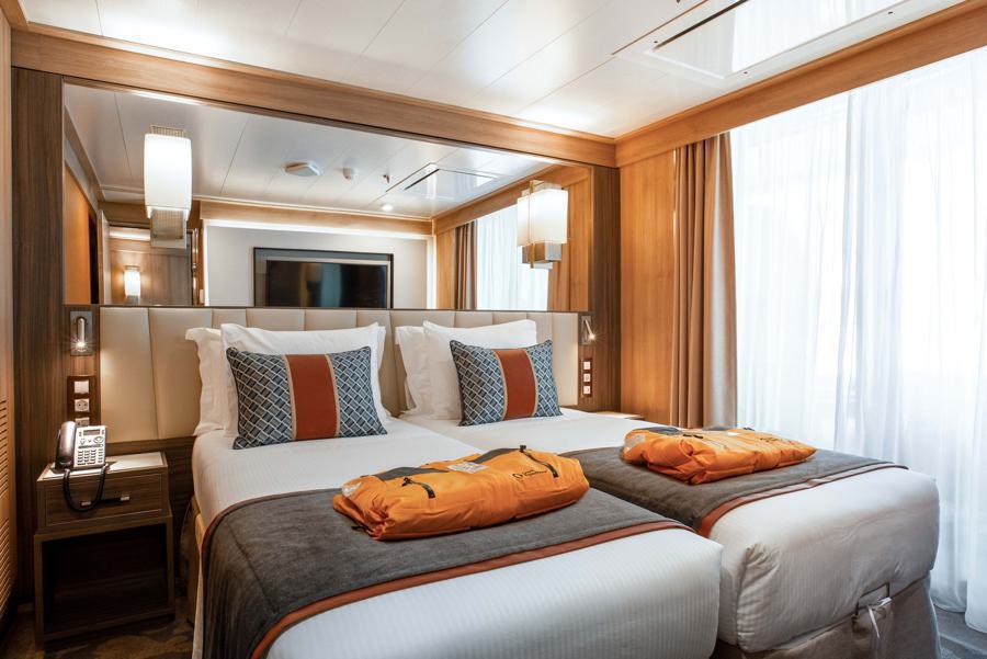 A modern, well-lit hotel room designed for the world explorer features two twin beds pushed together, each adorned with white linens and decorative pillows. Atop each bed is an orange robe. The room includes wooden furnishings, a flat-screen TV, and a large window with sheer curtains.