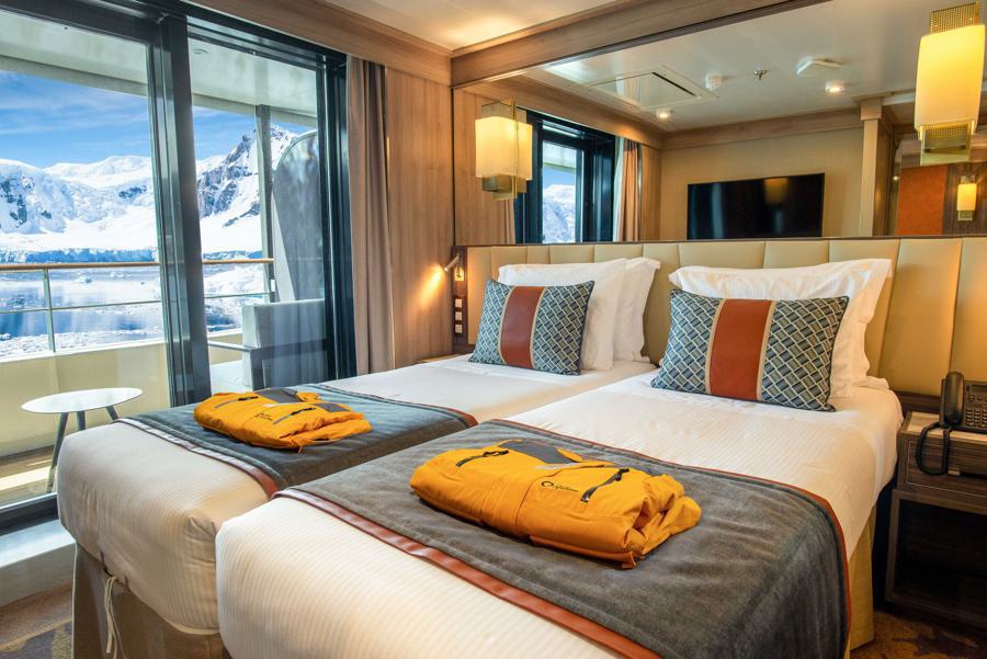 A well-furnished hotel room with two single beds topped with folded yellow robes. The room features a large window and glass door leading to a balcony with a scenic view of snowy mountains and a calm lake, perfect for the World Explorer on an Antarctica expedition. Warm lighting and modern decor complete the cozy ambiance.