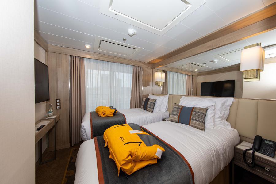 A well-lit hotel room features two twin beds with white linens and striped pillows. Each bed has a bright yellow life jacket placed on top. The room feels like part of a World Explorer Antarctica expedition, and includes a wall-mounted TV, a small desk, beige curtains, and warm, ambient lighting.