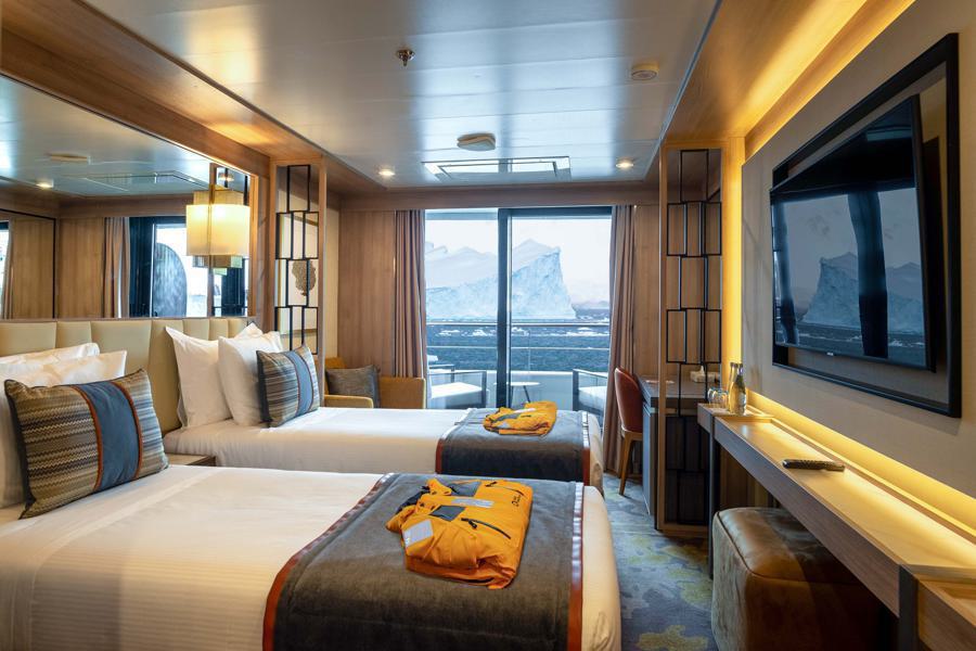 A cozy cruise ship cabin for World Explorer's Antarctica expedition features two neatly made beds with white and gray bedding and orange life jackets. The room has a large window revealing icebergs floating in the ocean. The cabin includes modern wooden furnishings, a flat-screen TV, and soft lighting.