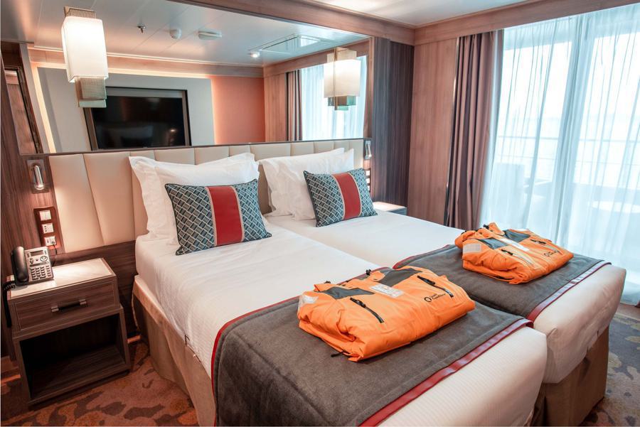 A modern cruise ship cabin features two neatly made twin beds pushed together, each topped with an orange life jacket. The room has a large window with sheer curtains, wooden accents, bedside tables with lamps, and a wall-mounted TV. Plush pillows add comfort to this World Explorer Antarctica expedition setting.