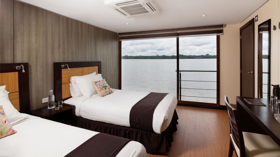 A hotel room with two neatly made twin beds featuring white linens and dark brown accents. A large window offers a scenic view of a river and forested area, similar to what you'd see on an Anakonda Amazon River Cruise ship. The room includes a desk, chair, and door leading to a private balcony. Modern lighting and air conditioning are present.