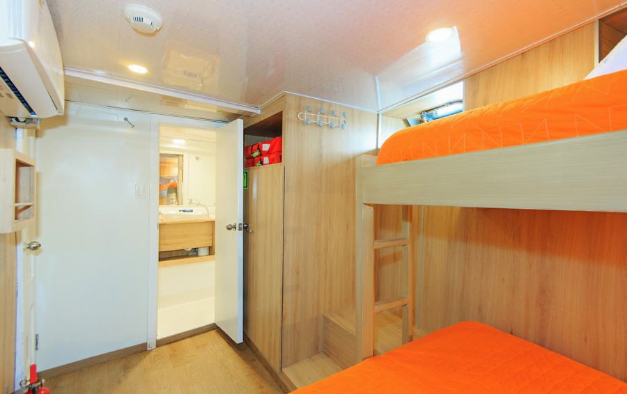 A compact room with wooden bunk beds, featuring wood paneling, bright orange bedding, and a small ladder for the top bunk. The door reveals a white and beige bathroom. Well-lit with ceiling lights, there’s also an air conditioner mounted on the wall—perfect for resting after an Aqua Galapagos cruise adventure.