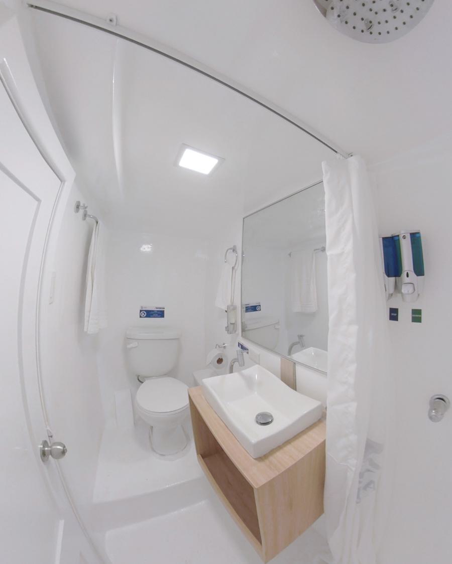 A modern, compact bathroom with a white toilet, a wall-mounted sink with a wooden cabinet beneath, and a large mirror above the sink. There is a shower area on the right with a white curtain and ceiling-mounted showerhead. The overall design is minimalistic, evoking the cleanliness of an Aqua Galapagos cruise.