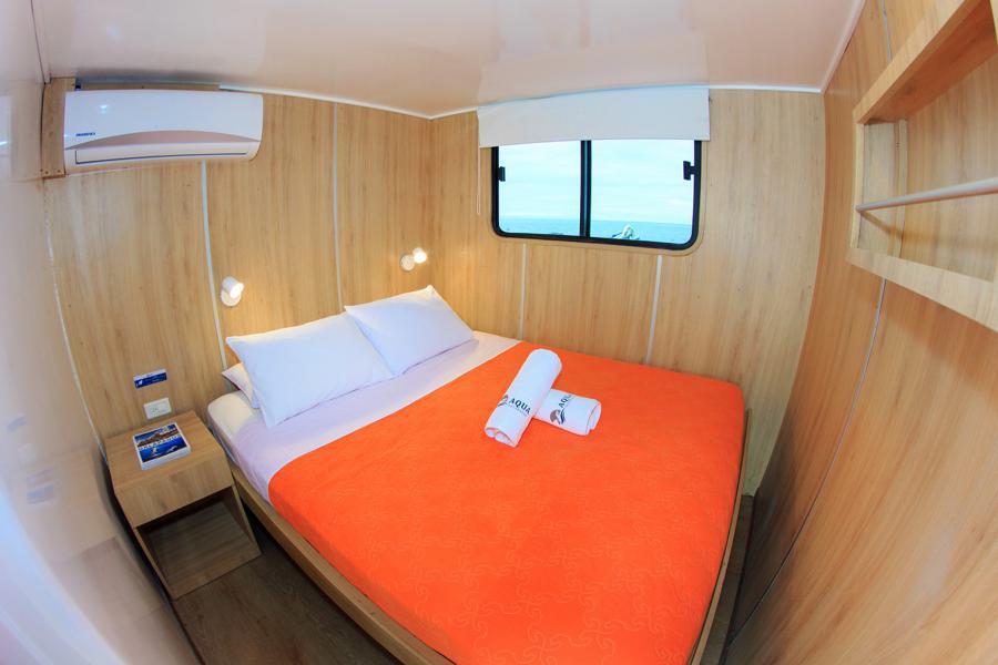 A cozy room with wooden walls features a double bed covered with an orange blanket, white pillows, and two rolled white towels on top. The room includes air conditioning, wall-mounted lamps, a small shelf, and a window overlooking the Aqua Galapagos cruise.