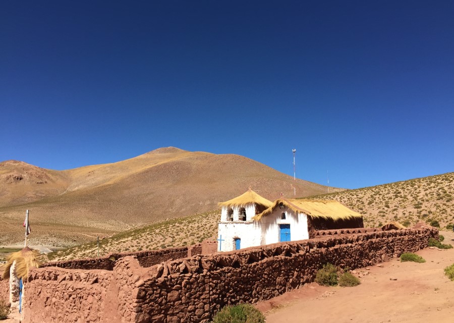 A small, traditional white church with a thatched roof stands against a backdrop of arid, rolling hills under a clear blue sky. Enclosed by a reddish-brown stone wall, this serene site in Chile's Atacama Desert features sparse bushes scattered throughout the dry landscape.