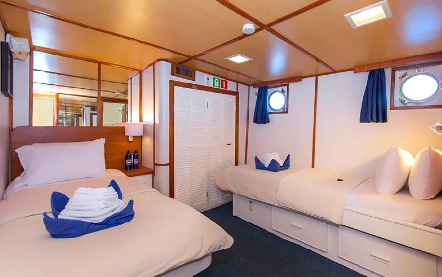 A cozy ship cabin with two neatly made single beds adorned with folded towels. The room features wood-paneled walls, blue curtains, round porthole windows, and a white closet. Soft lighting adds a warm ambiance to the clean and organized space, perfect for your Beluga Galapagos cruise.