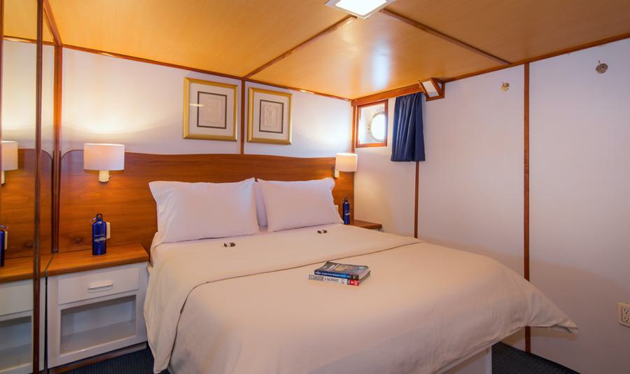 A cozy bedroom with wooden walls features a neatly made double bed with white linens and pillows. There are two framed pictures above the bed, two bedside tables with lamps, a small round window with a blue curtain, and a book placed on the bed, evoking the comfort of a Beluga Galapagos cruise.